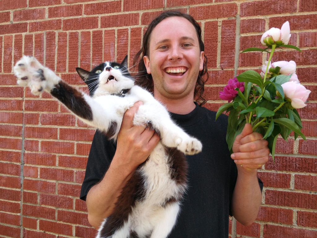 The artist, Dylan Cale Jones, stands, laughing, in front of a brick wall. In their left hand, they hold a fresh bunch of full, pink peonies. In their right hand they hold an angry cat. The cat hisses and swipes at the photographer.
