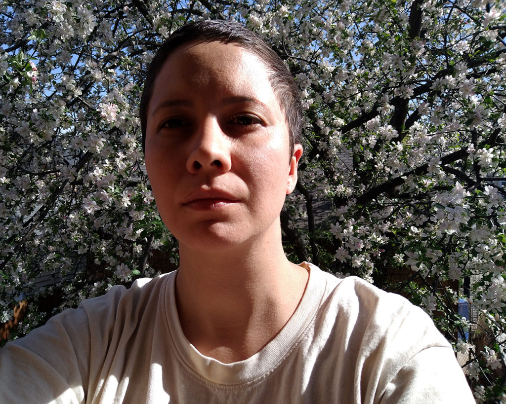 The artist, Isa Rodriguez, stands in front of an apple tree in full bloom. The cloud of blossoms fills the space behind them. The sun shines warmly on the left side of their face and left shoulder.