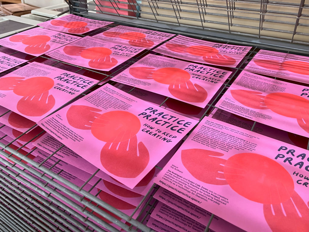 Freshly printed pages are arranged neatly on a large metal drying rack. Each page shows an illustration of two hands cradling a glowing orb gently on their fingertips. The legible text reads, "Practice Practice: How to Keep Creating"