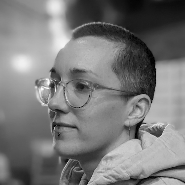 Black and white portrait of KT Duffy. They have a shaved head, a lip piercing, glasses and are wearing a hoodie. The background is out of focus. 