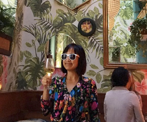 Denise Duong is wearing sunglasses, smiling and holding up a glass of wine. She is standing in a room decorated with wallpaper printed with tropical plants. 