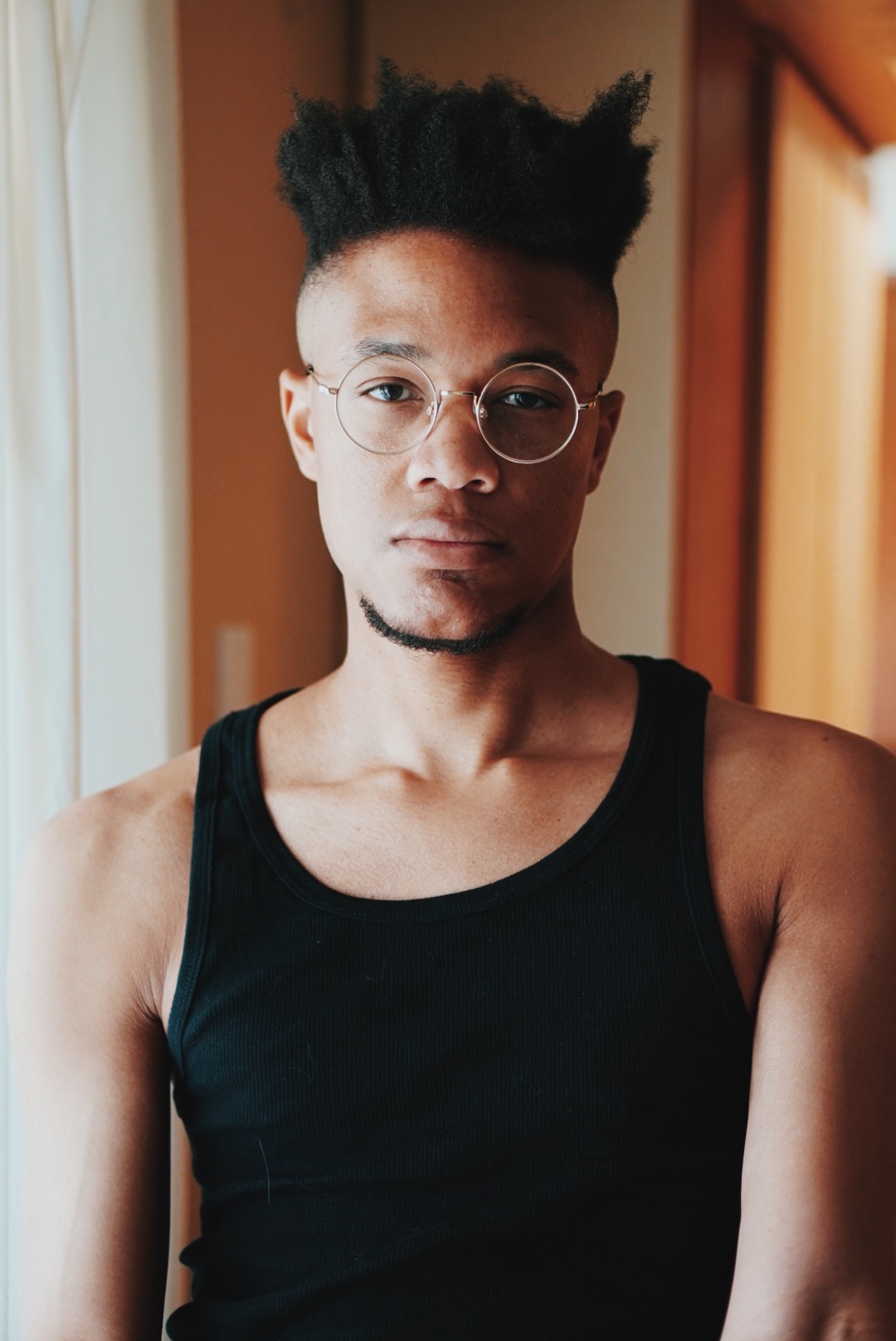 Portrait of artist, Black Masq. He is looking directly in the camera. He is wearing a black tank top and round wire frame glasses.