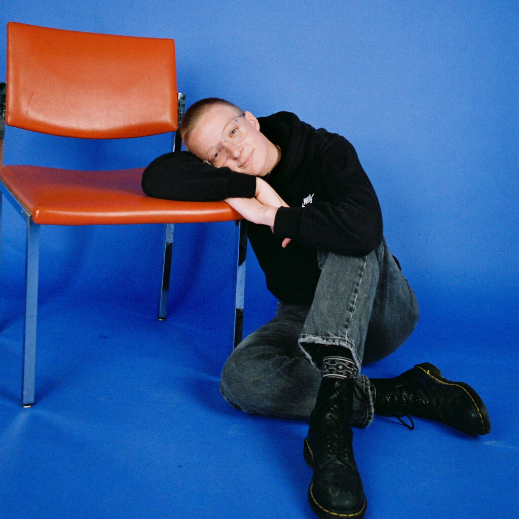 Portrait of artist Erin Owen. They are sitting on the floor, leaning against an orange chair and smiling. 