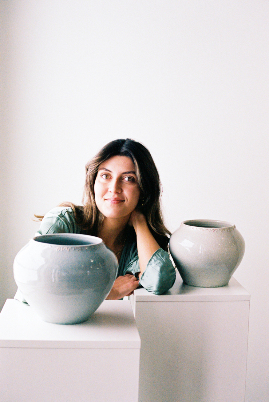 Portrait of artist Amy Sanders de Melo. She is sitting in between 2 of her vases. Her elbow is resting on a pedestal and she is smiling.
