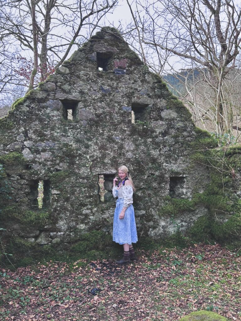 Artist Martha Coates Donahoo stands outside, in front of a dilapidated stone cottage.