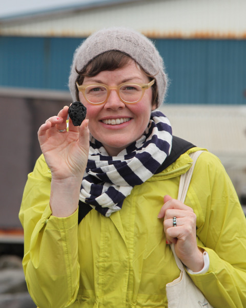 Portrait of artist M Wright. M is wearing a yellow rain jacket, a black and white stripped scarf and a grey beanie. She is holding up a rock and smiling brightly. 